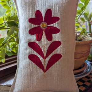 May include: A rectangular off-white fabric sachet featuring a centered embroidered flower with deep red petals, a yellow center, and a thin gold stem. A wooden ruler at the bottom shows the item measures approximately 7 inches in width.
