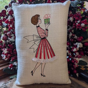 May include: A rectangular off-white fabric cushion featuring an embroidered illustration of a person with brown hair wearing a red and white patterned dress with a large bow, holding a green gift box with a red bow, positioned above a ruler.