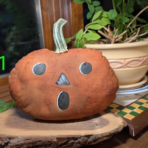 May include: A plush pumpkin decoration with a textured brown fabric surface, a green stem, and a hand-painted face featuring two round eyes, a triangular nose, and an oval mouth. The number #1 is printed in bright green on the left side.