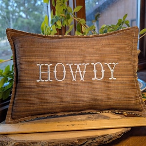 May include: Brown rectangular pillow with the embroidered word "HOWDY" in white. The pillow has a striped pattern and a decorative border. The pillow is resting on a wooden surface.