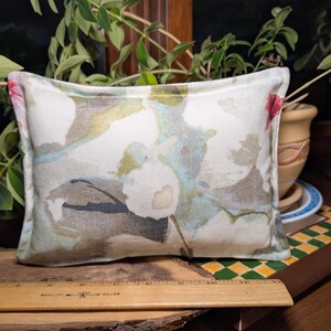 May include: Rectangular decorative pillow with a floral pattern in shades of white, pink, blue, and green. The pillow is placed on a wooden surface with a ruler. The background includes plants and a ceramic pot.