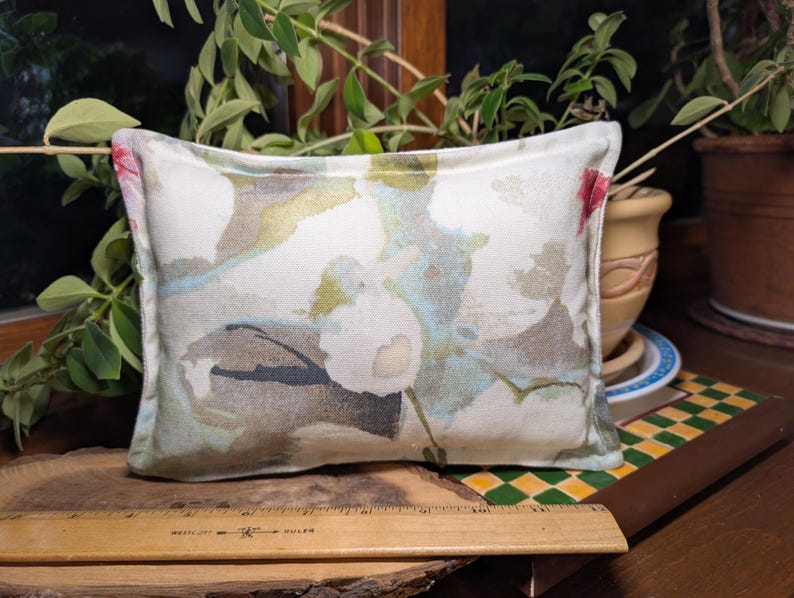 May include: Rectangular decorative pillow with a floral pattern in shades of white, pink, blue, and green. The pillow is placed on a wooden surface with a ruler. The background includes plants and a ceramic pot.