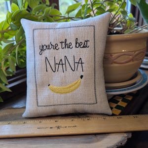 May include: A small, square, off-white fabric cushion featuring black embroidered text that reads 'you're the best NANA' above a yellow banana graphic. A wooden ruler is placed in front, showing the cushion is approximately 12.7 cm wide.