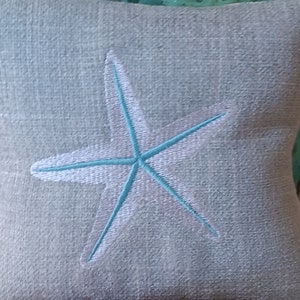 Peut inclure: Un coussin carr&eacute; bleu clair avec une &eacute;toile de mer brod&eacute;e. L'&eacute;toile de mer est bord&eacute;e de blanc et remplie de bleu clair. Le coussin a un aspect textur&eacute;, probablement en lin ou dans un tissu similaire.