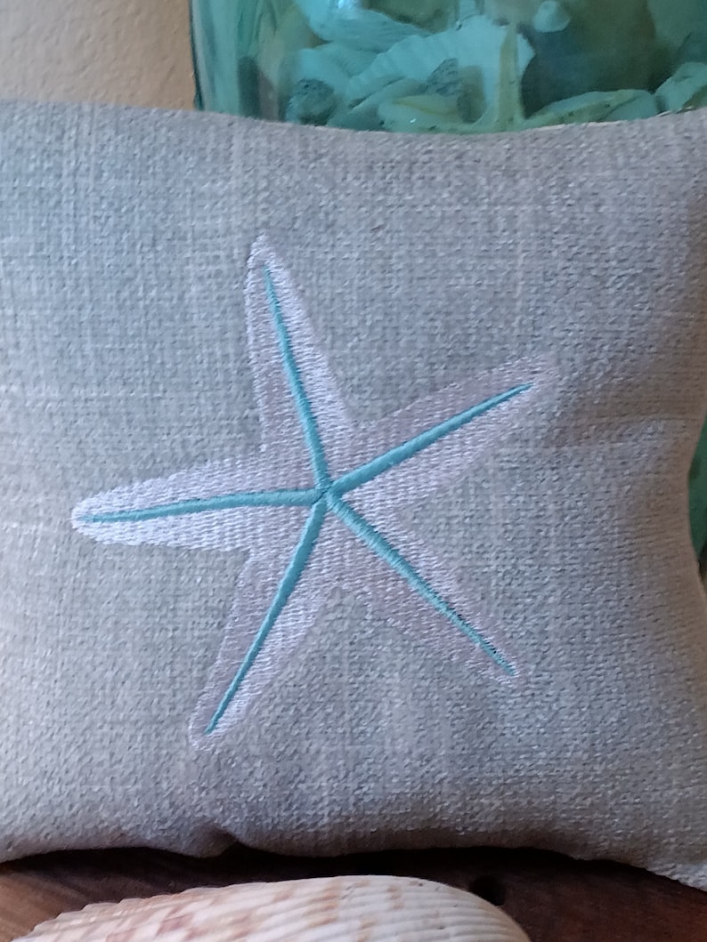 Peut inclure: Un coussin carr&eacute; bleu clair avec une &eacute;toile de mer brod&eacute;e. L'&eacute;toile de mer est bord&eacute;e de blanc et remplie de bleu clair. Le coussin a un aspect textur&eacute;, probablement en lin ou dans un tissu similaire.