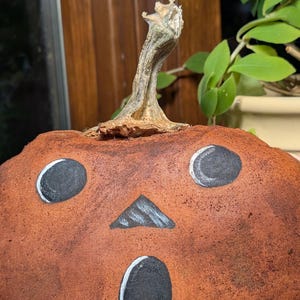 May include: A fabric pumpkin decoration with a textured, mottled brown surface. It features a painted face with two round black eyes, a triangular black nose, and an oval black mouth, all outlined in white. A dried, woody stem sits on top.