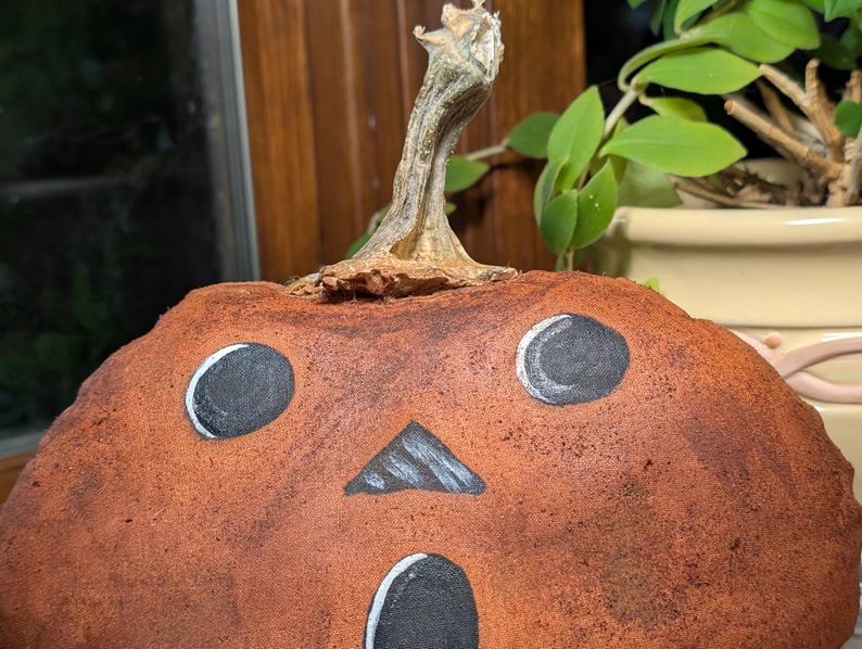 May include: A fabric pumpkin decoration with a textured, mottled brown surface. It features a painted face with two round black eyes, a triangular black nose, and an oval black mouth, all outlined in white. A dried, woody stem sits on top.