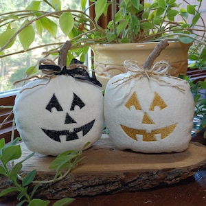 May include: Two white fabric pumpkins with carved jack-o'-lantern faces. One has a black face, the other a gold face. Both have twig stems and are decorated with twine and ribbon. They sit on a wooden slice, perfect for Halloween decor.