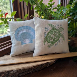 Peut inclure: Deux petits coussins carr&eacute;s d&eacute;coratifs blanc cass&eacute;. L'un pr&eacute;sente un motif de coquillage bleu clair, l'autre une tortue verte avec des bulles. Une r&egrave;gle en bois est pos&eacute;e devant les coussins.