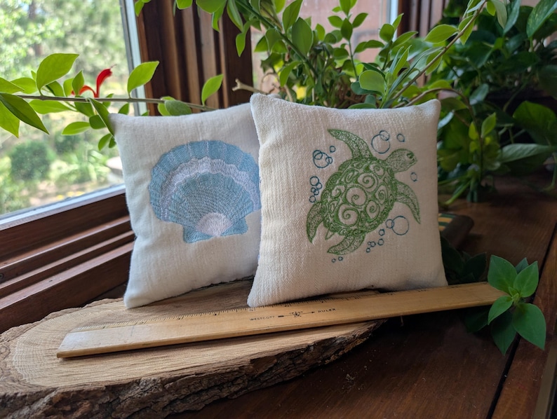 Peut inclure: Deux petits coussins carr&eacute;s d&eacute;coratifs blanc cass&eacute;. L'un pr&eacute;sente un motif de coquillage bleu clair, l'autre une tortue verte avec des bulles. Une r&egrave;gle en bois est pos&eacute;e devant les coussins.