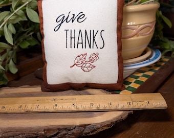 May include: A small square fabric pillow with a white center and brown border, featuring the black text "give THANKS" and a red embroidered leaf design. A wooden ruler placed in front shows the pillow is approximately 8 inches wide.