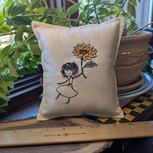 May include: A small, square, off-white fabric sachet featuring an embroidered design of a person with curly hair holding a large yellow sunflower. A wooden ruler placed in front shows the item measures approximately 5 inches in width.