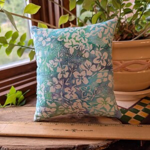Peut inclure: Un petit coussin carr&eacute; avec un motif floral dans des tons de bleu et blanc. Le coussin est pos&eacute; sur une surface en bois avec une r&egrave;gle. L'arri&egrave;re-plan comprend de la verdure et un pot d&eacute;coratif.
