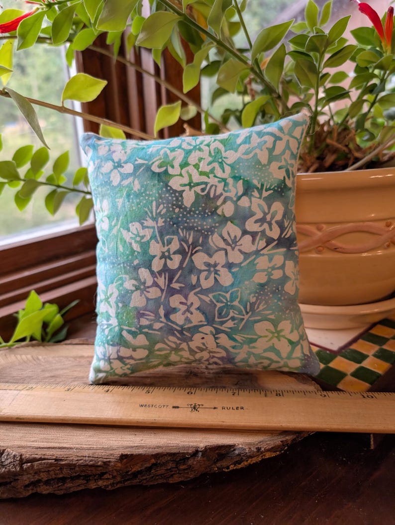 Peut inclure: Un petit coussin carr&eacute; avec un motif floral dans des tons de bleu et blanc. Le coussin est pos&eacute; sur une surface en bois avec une r&egrave;gle. L'arri&egrave;re-plan comprend de la verdure et un pot d&eacute;coratif.