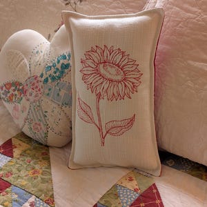 May include: A rectangular cream-coloured throw pillow featuring a single red embroidered sunflower design on the front. The pillow has a piped edge and a textured fabric finish, standing upright against a quilted fabric surface with floral patterns.