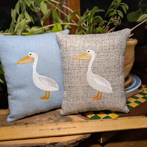 Peut inclure: Deux coussins d&eacute;coratifs carr&eacute;s. L'un est bleu clair, l'autre beige. Chaque coussin pr&eacute;sente un p&eacute;lican blanc brod&eacute; avec un bec et des pieds jaunes. Les coussins sont en tissu textur&eacute;.