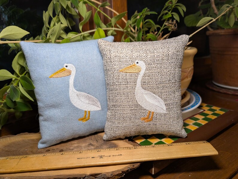 Peut inclure: Deux coussins d&eacute;coratifs carr&eacute;s. L'un est bleu clair, l'autre beige. Chaque coussin pr&eacute;sente un p&eacute;lican blanc brod&eacute; avec un bec et des pieds jaunes. Les coussins sont en tissu textur&eacute;.