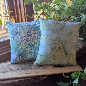 Peut inclure: Deux petits coussins carr&eacute;s en tissu. L'un est bleu clair avec un motif floral blanc. L'autre est vert clair avec un motif mouchet&eacute;. Ils reposent sur une surface en bois pr&egrave;s d'une fen&ecirc;tre avec de la verdure.