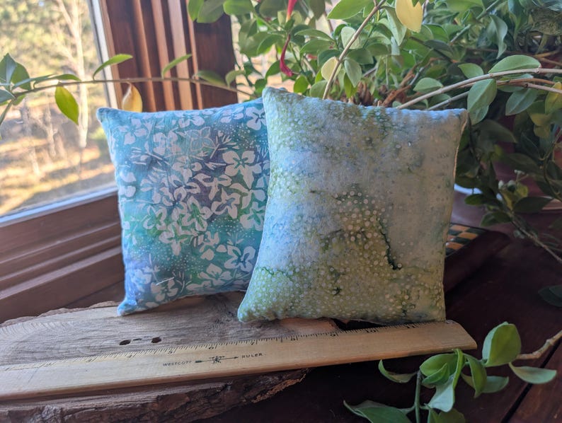 Peut inclure: Deux petits coussins carr&eacute;s en tissu. L'un est bleu clair avec un motif floral blanc. L'autre est vert clair avec un motif mouchet&eacute;. Ils reposent sur une surface en bois pr&egrave;s d'une fen&ecirc;tre avec de la verdure.