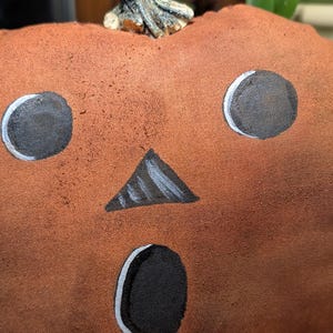 May include: A plush pumpkin decoration made of textured orange fabric with a painted face featuring two round black eyes, a triangular black nose, and an oval black mouth, topped with a small, realistic-looking brown stem.