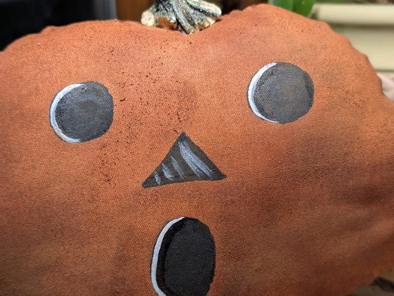 May include: A plush pumpkin decoration made of textured orange fabric with a painted face featuring two round black eyes, a triangular black nose, and an oval black mouth, topped with a small, realistic-looking brown stem.