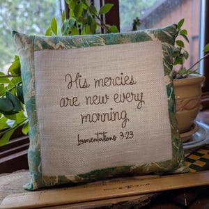 May include: Square decorative pillow with a white center and a green patterned border. The pillow features embroidered text that reads "His mercies are new every morning, Lamentations 3:23". The pillow is placed on a wooden surface.