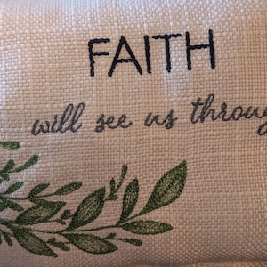 May include: Cream-colored fabric with embroidered text reading "FAITH will see us through." The text is in dark gray and black. A green embroidered leaf design is in the lower left corner.