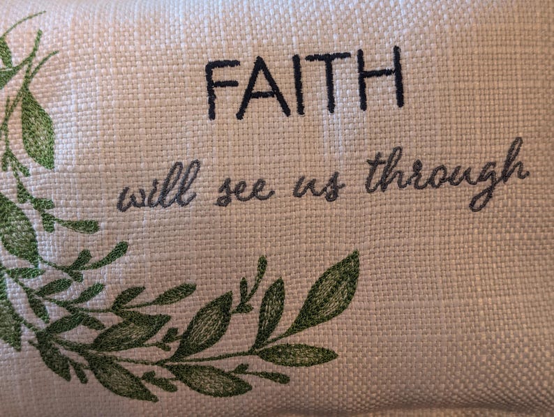 May include: Cream-colored fabric with embroidered text reading "FAITH will see us through." The text is in dark gray and black. A green embroidered leaf design is in the lower left corner.
