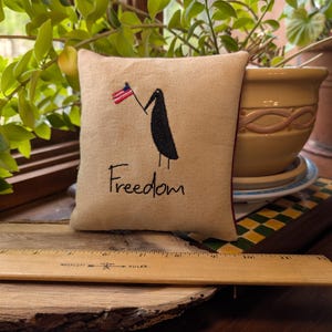 May include: A small, beige pillow with a black bird illustration holding a small American flag. The word "Freedom" is written in black script below the bird. The pillow has a red border.