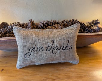 May include: A rectangular burlap pillow with the words "give thanks" embroidered in black script. The pillow is resting on a wooden surface, with a wooden bowl filled with pine cones in the background.