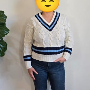 May include: Cream-colored cable knit sweater with a V-neck and blue and navy stripes. The sweater has long sleeves and a relaxed fit. The person is wearing blue jeans.
