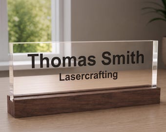 Op de afbeelding: Een rechthoekig bureau-naamplaatje van helder acryl met de zwart gedrukte tekst Thomas Smith en Lasercrafting, stevig geplaatst in een donkerbruine houten voet. Het bord is horizontaal geplaatst en heeft een strak, transparant ontwerp.