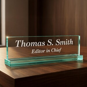 Op de afbeelding: Een bureau-naamplaatje van helder glas met een rechthoekige voet en een verticaal paneel. De tekst "Thomas S. Smith" is in een schreeflettertype gegraveerd, met "Editor in Chief" in een kleiner schreeflettertype op de regel eronder.