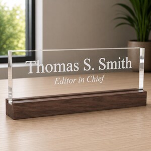Op de afbeelding: Een rechthoekig bureau-naamplaatje van helder acryl met de gegraveerde witte tekst Thomas S. Smith en Editor in Chief, geplaatst in een massief donkerbruine houten voet met een horizontale groef voor extra stabiliteit.