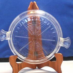 May include: A clear glass pie plate with a raised, circular design in the center and two handles on the sides. The plate is on a wooden stand.