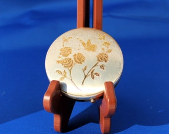 May include: A silver compact mirror with a floral design featuring gold roses and a butterfly. The mirror is on a wooden stand.