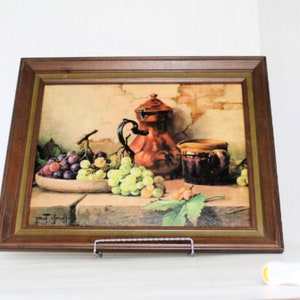 May include: A framed still life painting of a red pitcher, grapes, and a jar. The painting is set against a background of a brick wall.