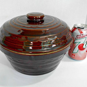 Pode incluir: Um prato de cerâmica marrom esmaltado com tampa. O prato tem um design ondulado e uma borda dourada. O prato está ao lado de uma lata de 7 Up de cereja dietética.