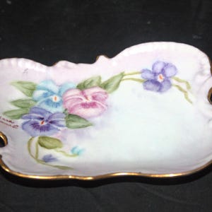 May include: A small, white porcelain dish with a scalloped edge and a gold trim. The dish is decorated with hand-painted purple, blue, and pink pansies. The signature "Linda Schmidt" is visible on the bottom left side of the dish.