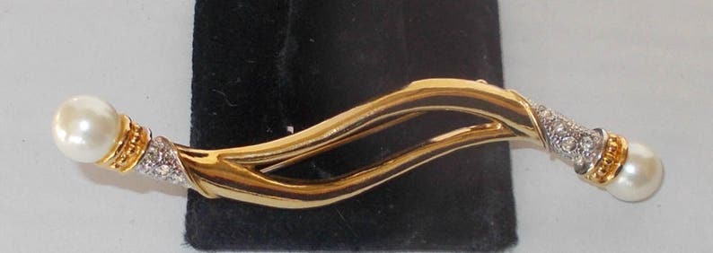 Pu&ograve; includere: Un fermaglio dorato con un design ondulato e due accenti di perle finte.