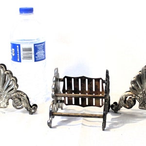 May include: Three silver-toned metal napkin holders. Two are ornate with a shell-like design and the third is a simple rectangular design with a decorative border.