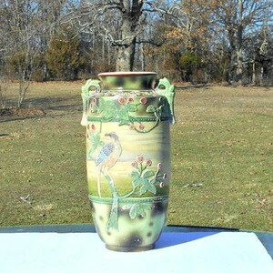 May include: A tall ceramic vase featuring a light tan body with green and brown gradient accents. The surface is decorated with a raised relief design of a long-tailed bird perched among green leaves and pink flowers, with two handles.