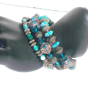May include: A set of four bracelets featuring turquoise and green glass beads, copper toned metal beads, and decorative metal charms. The bracelets are layered on a black mannequin arm.