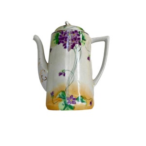 Puede incluir: Una tetera de cerámica blanca con un diseño floral. La tetera tiene una base amarilla, naranja y verde con flores moradas y hojas verdes. La tetera tiene un borde dorado.