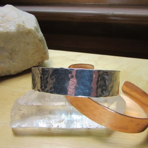May include: Two bracelets, one silver and one copper, both with a hammered texture. The bracelets are stacked on top of each other.