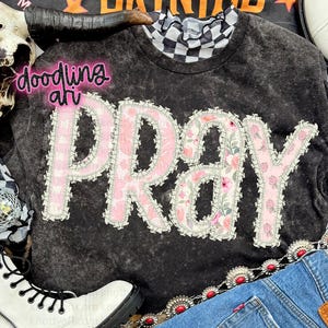 May include: Dark grey sweatshirt featuring the word "PRAY" in a floral design with pink scalloped edging. The sweatshirt has a black and white checkered collar. The text "doodling art" is also visible.