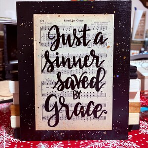 May include: A framed artwork with a black border and the text "Just a Sinner Saved by Grace" in elegant script. The background features sheet music, with gold and silver glitter accents. The artwork is displayed on a red and white snowflake patterned surface.
