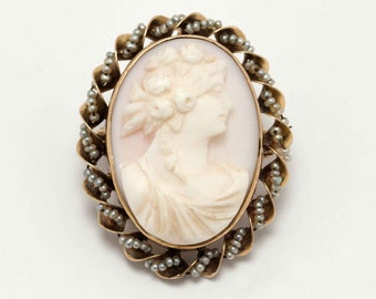 Puede incluir: Broche de camafeo ovalado con un perfil tallado de mujer en relieve crema sobre un fondo rosa pálido. El marco dorado está adornado con pequeñas cuentas grises y un borde decorativo en forma de hoja. Una joya clásica.