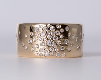 May include: A wide gold band ring featuring a cluster of numerous round, clear, brilliant-cut diamonds set flush into the surface. The diamonds vary in size and are arranged in a dense, scattered pattern across the front of the polished gold metal.