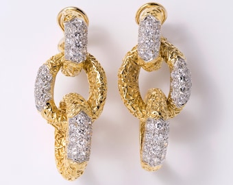 May include: A pair of drop earrings featuring two interlocking oval links with a textured gold finish. Each link is accented with sections of pavé-set clear stones in a silver-toned setting, creating a layered, dangling design.